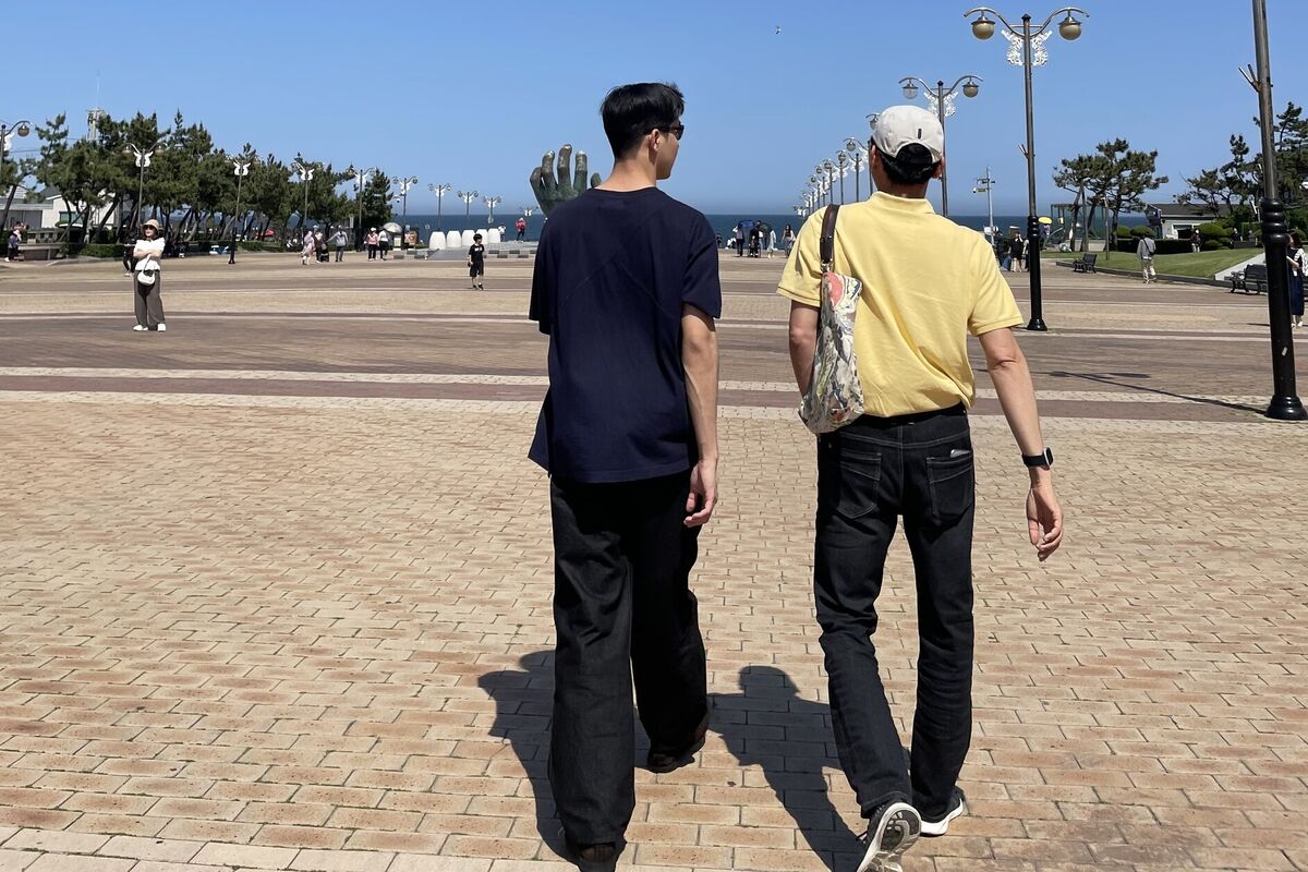 Rok and a guest walking together across a seaside plaza, talking, seen from behind